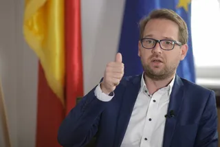 Un bărbat cu ochelari și sacou albastru stă în fața unor steaguri și face gestul cu degetul mare ridicat.