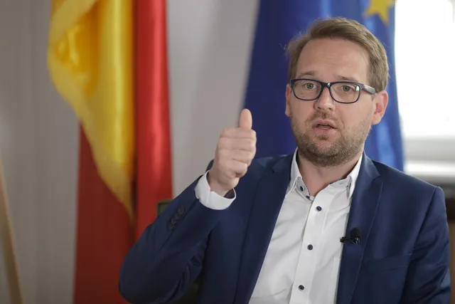 Un bărbat cu ochelari și sacou albastru stă în fața unor steaguri și face gestul cu degetul mare ridicat.