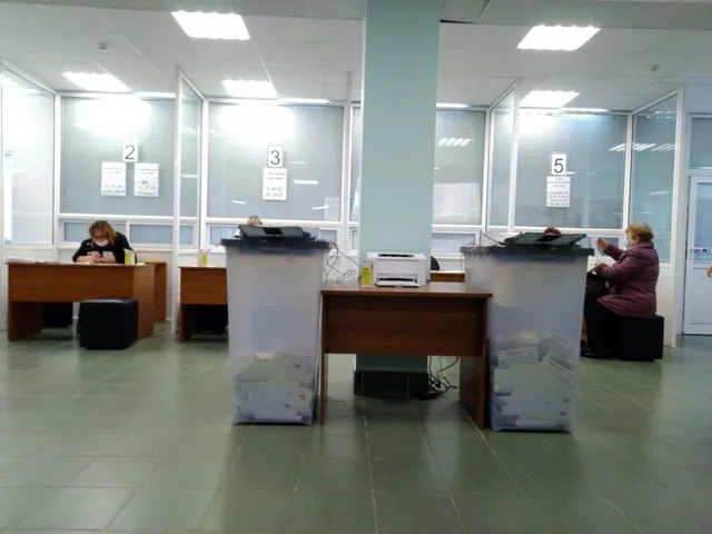 O persoană stă la un birou în fața unei cabine de vot, lângă un dispozitiv alb și un suport gri cu hârtii.