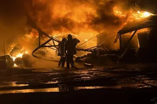 Două siluete de pompieri stau în fața unei structuri metalice arse și a unor flăcări mari într-un mediu întunecat și umed.