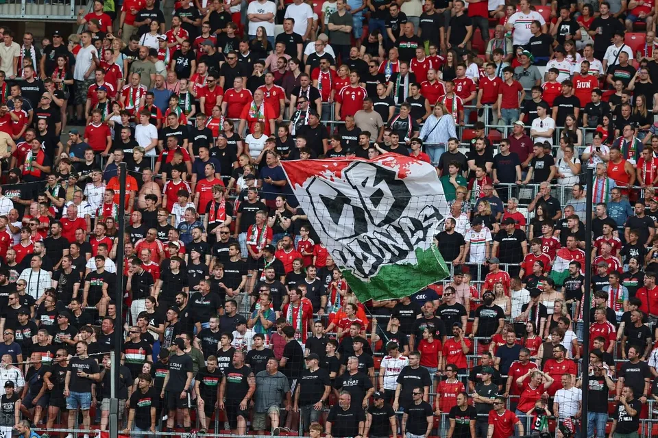 O mulțime de oameni stă pe tribunele unui stadion, mulți purtând haine roșii, în timp ce un grup ridică un steag mare cu un simbol central.