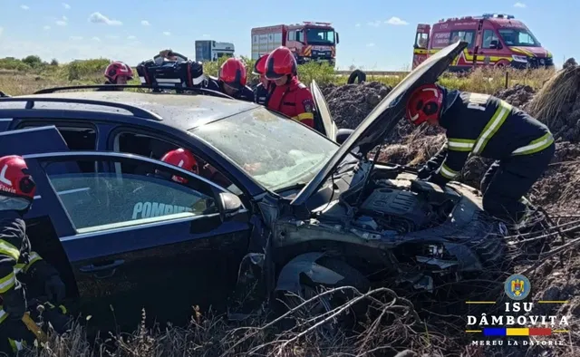 Mai mulți salvatori cu echipament de protecție roșu și negru lucrează în jurul unui autoturism negru avariat situat într-un teren cu vegetație uscată.