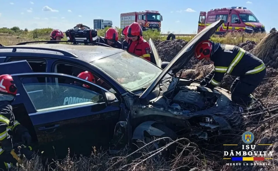 Mai mulți salvatori cu echipament de protecție roșu și negru lucrează în jurul unui autoturism negru avariat situat într-un teren cu vegetație uscată.