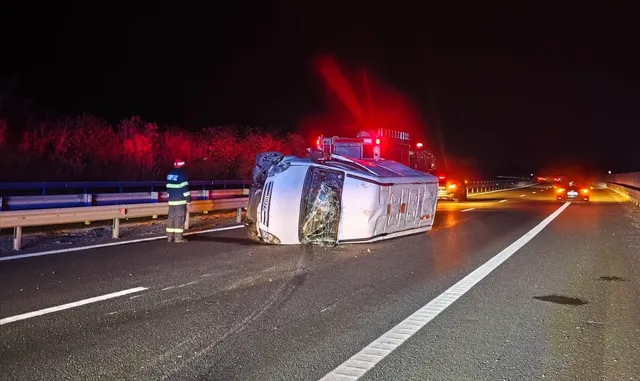 O autoutilitară albă este răsturnată pe o laterală pe o autostradă, lângă un gard de protecție, sub lumini roșii de semnalizare nocturnă.