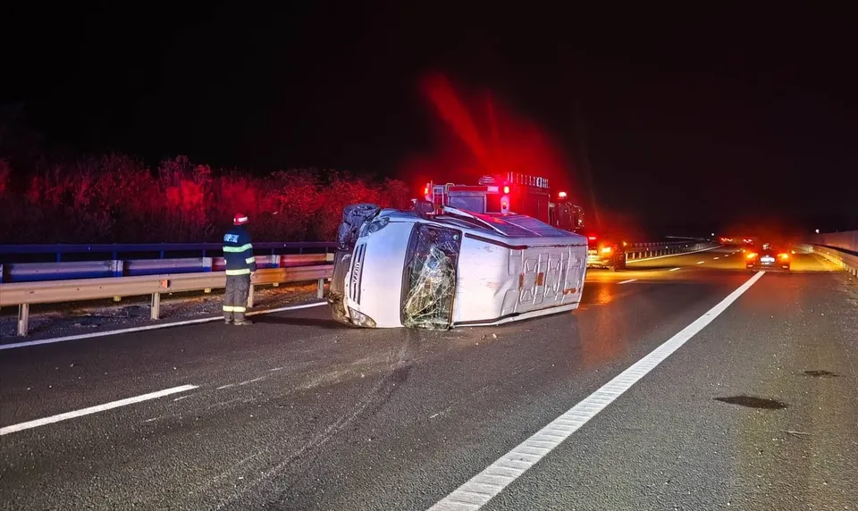 O autoutilitară albă este răsturnată pe o laterală pe o autostradă, lângă un gard de protecție, sub lumini roșii de semnalizare nocturnă.
