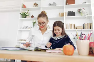 O femeie cu ochelari și o copilă stau la o masă de lemn, scriind în cărți de lângă un recipient cu instrumente de scris colorate.