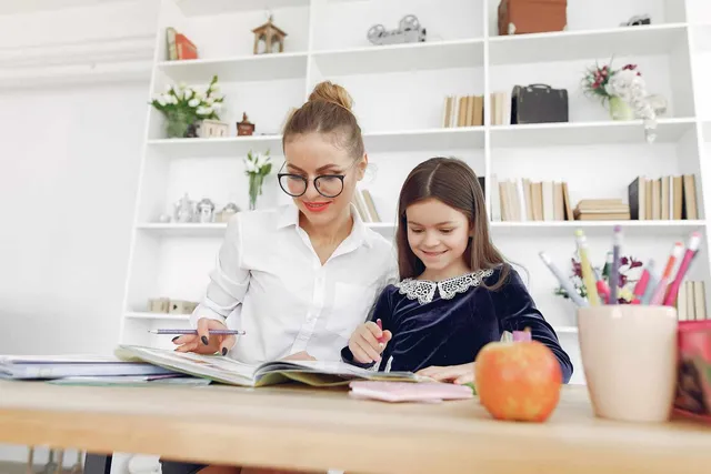 O femeie cu ochelari și o copilă stau la o masă de lemn, scriind în cărți de lângă un recipient cu instrumente de scris colorate.