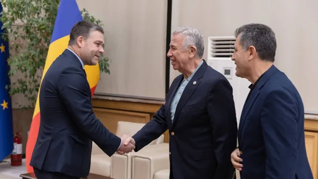 Un bărbat într-un costum închis se strânge pe mână cu un alt bărbat într-un costum închis, în timp ce un al treilea bărbat îi privește.