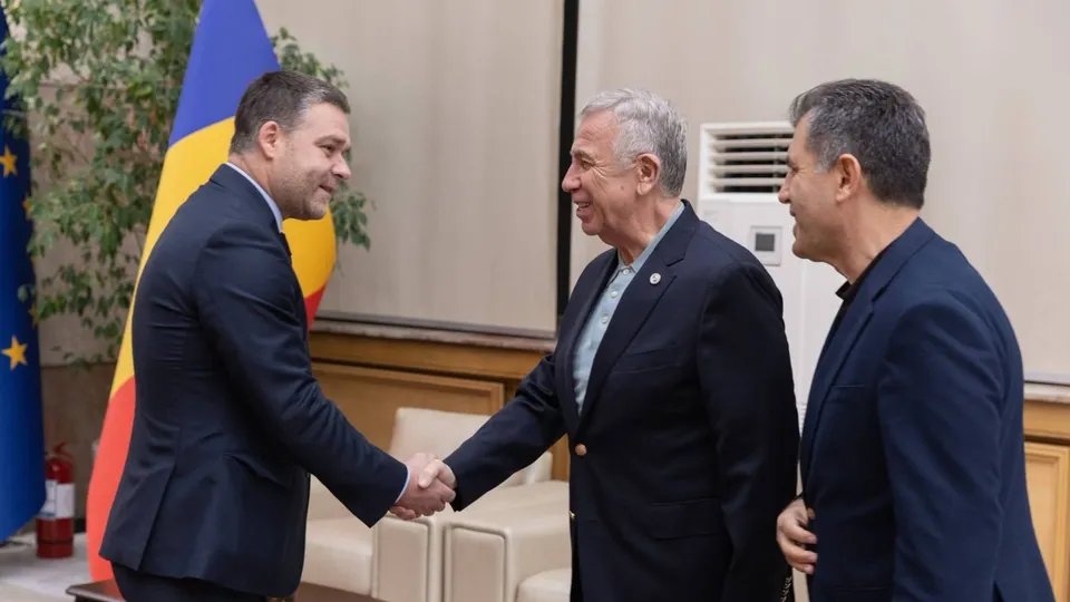 Un bărbat într-un costum închis se strânge pe mână cu un alt bărbat într-un costum închis, în timp ce un al treilea bărbat îi privește.