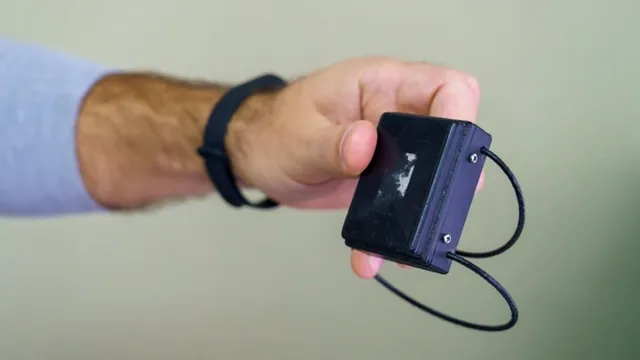 O mână care poartă o brățară neagră în jurul încheieturii ține un dispozitiv electronic dreptunghiular negru cu un cablu subțire atașat de acesta.