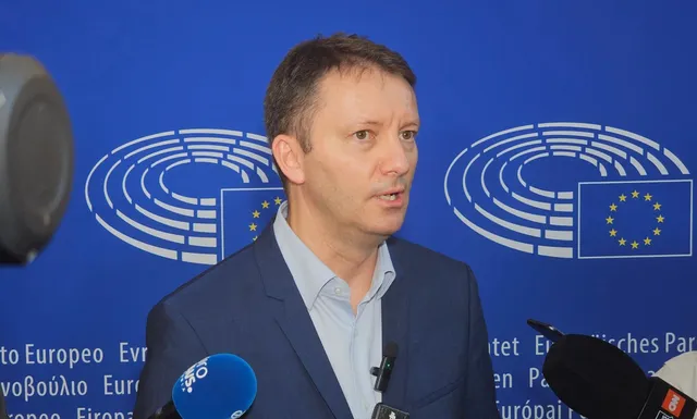 Un bărbat cu părul scurt, purtând sacou albastru și cămașă deschisă, stă în fața unui fundal albastru cu embleme circulare albe.