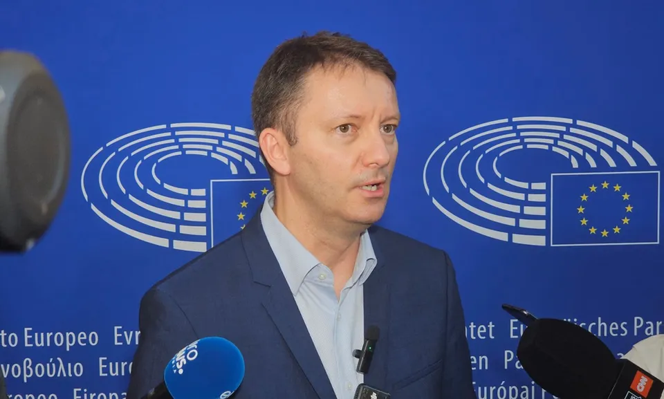 Un bărbat cu cămașă deschisă și sacou stă în fața unui fundal albastru cu simboluri grafice albe și microfoane în prim-plan.