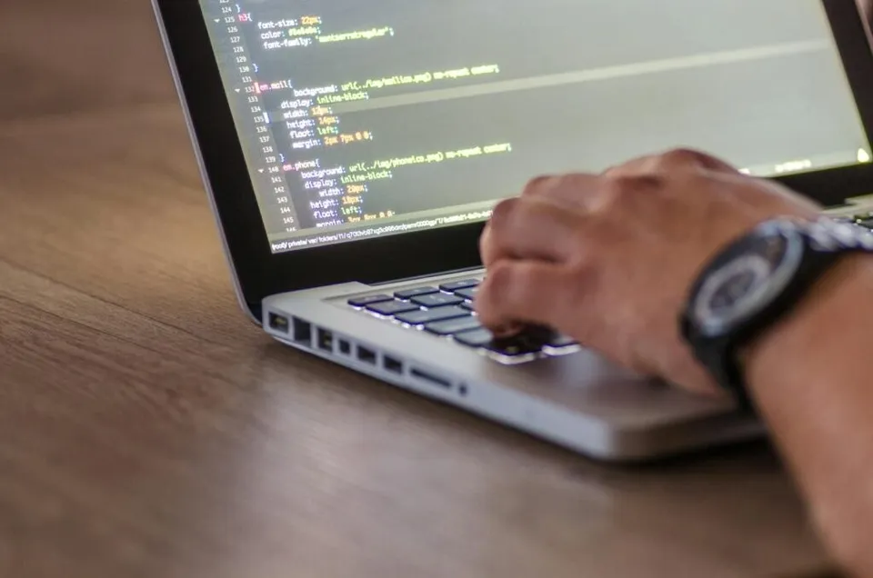 Mâinile unei persoane tastând pe un laptop deschis aflat pe o suprafață de lemn.