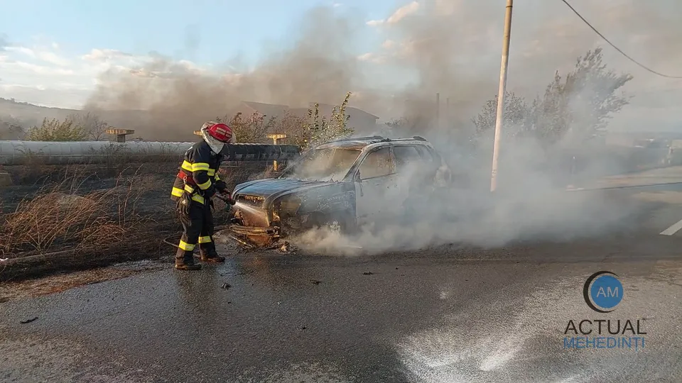 Un pompier în echipament de protecție stă lângă un autoturism acoperit de fum pe un drum asfaltat.