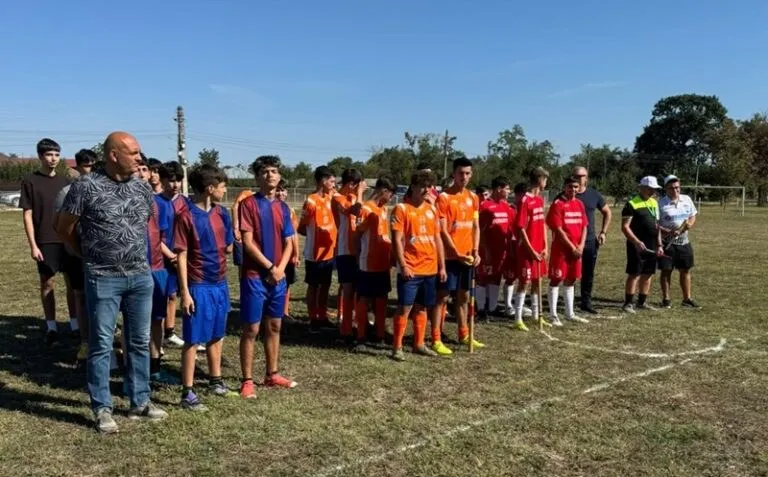 Un bărbat în blugi și mai multe grupuri de tineri în tricouri sportive stau pe o peluză, lângă linii albe.