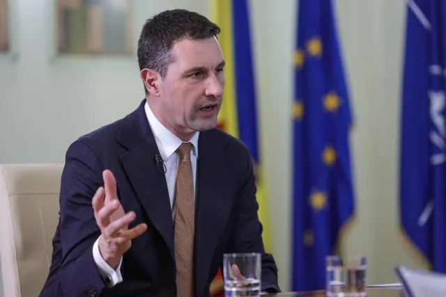 Un bărbat cu păr scurt, purtând sacou închis, cămașă albă și cravată maro, stă la o masă cu pahare de sticlă în față.