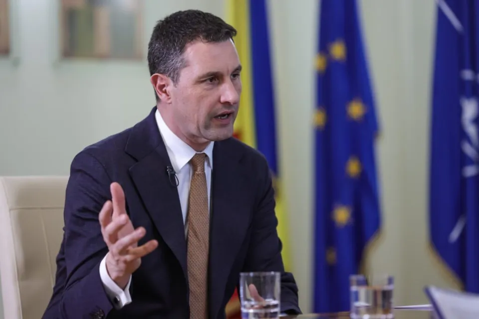 Un bărbat cu păr scurt, purtând sacou închis, cămașă albă și cravată maro, stă la o masă cu pahare de sticlă în față.