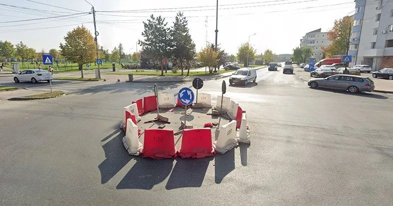 Un cerc de bariere de plastic roșii și albe delimitează o zonă pe asfaltul unei intersecții urbane sub un cer senin.