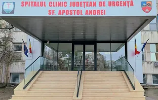 Intrarea unui spital este marcată de un panou roșu cu text alb, având scări cu balustrade de sticlă și două steaguri pe lateral.