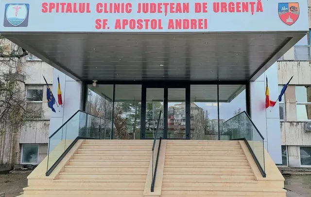 Intrarea unui spital este marcată de un panou roșu cu text alb, având scări cu balustrade de sticlă și două steaguri pe lateral.