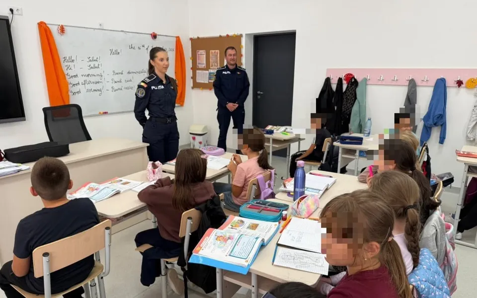 Două persoane în uniforme de poliție stau în picioare într-o sală de clasă în fața unor elevi așezați la bănci cu caiete și rechizite.