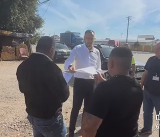 Un bărbat într-o cămașă albă ține o foaie de hârtie în timp ce discută cu alți trei bărbați pe o stradă cu mașini și un camion.