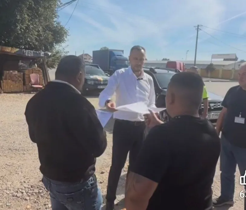 Un bărbat într-o cămașă albă ține o foaie de hârtie în timp ce discută cu alți trei bărbați pe o stradă cu mașini și un camion.