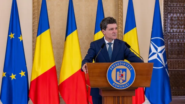 Un bărbat într-un costum închis la culoare și cravată stă în spatele unui podium de lemn în fața mai multor steaguri.