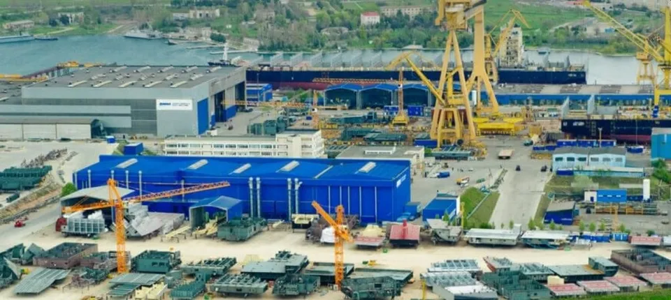 O vedere aeriană a unui șantier naval cu macarale galbene, clădiri industriale gri și albastre și diverse structuri metalice pe un teren vast.