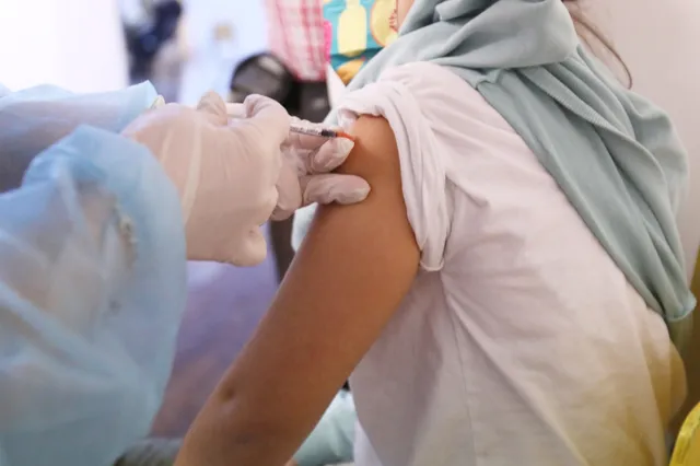 O persoană cu mâini înfășurate în mănuși albe primește o injecție în brațul drept de la o persoană purtând halat albastru și mănuși medicale.