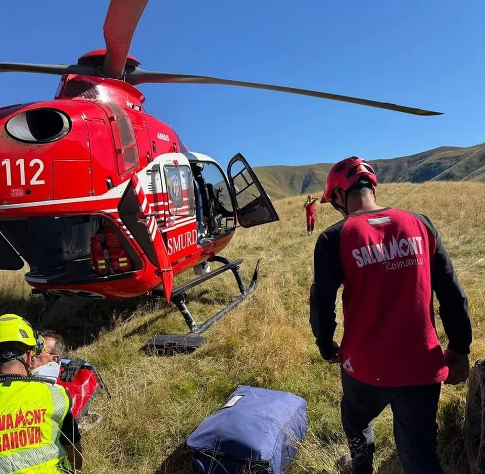 Un elicopter roșu cu inscripția 112 stă pe o iarbă verde lângă un salvamontist care poartă o vestă roșie cu textul Salvamont România.