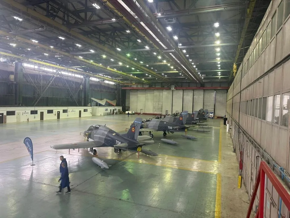 Mai multe avioane de culoare închisă sunt parcate într-un hangar mare cu lumină artificială, în timp ce un bărbat merge pe podeaua verde.