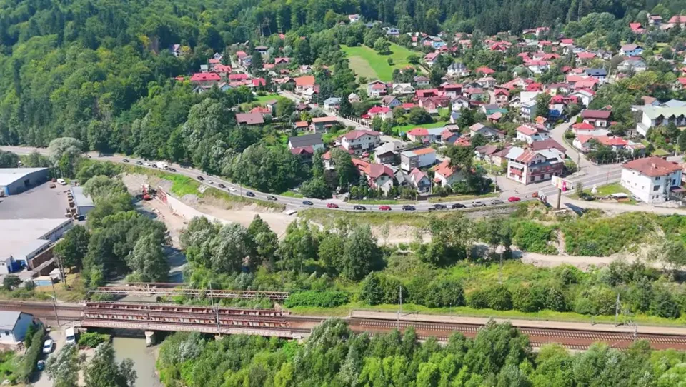 O vedere aeriană prezintă o zonă rezidențială cu case cu acoperișuri colorate, o șosea sinuoasă și o cale ferată traversând un peisaj împădurit.