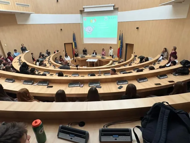 O grupă de persoane este așezată la mese curbe în fața unui ecran de proiecție care afișează un fundal verde cu text și grafice.