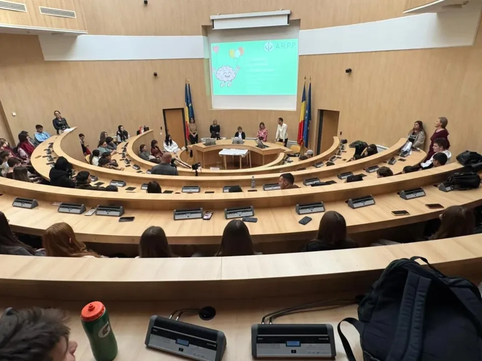 O grupă de persoane este așezată la mese curbe în fața unui ecran de proiecție care afișează un fundal verde cu text și grafice.
