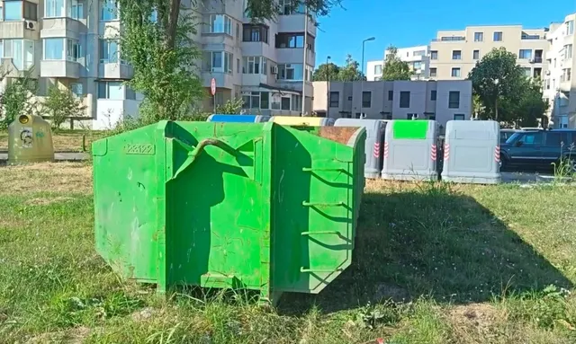 Un container mare de culoare verde este situat pe o suprafață cu iarbă, în fața unor clădiri de locuințe și a altor containere de deșeuri.