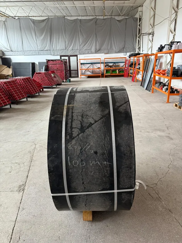 Un rulou mare de material negru este așezat pe un suport de lemn într-o hală industrială cu rafturi și utilaje în fundal.