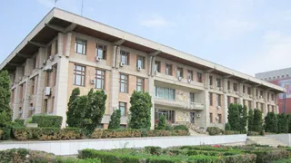 O clădire înaltă de beton cu ferestre multiple, înconjurată de garduri vii și copaci verzi sub un cer senin.