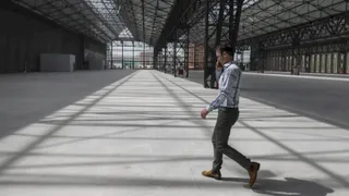 Un bărbat într-un tricou cu mânecă lungă și pantaloni gri merge printr-un spațiu industrial mare și gol cu structură metalică.