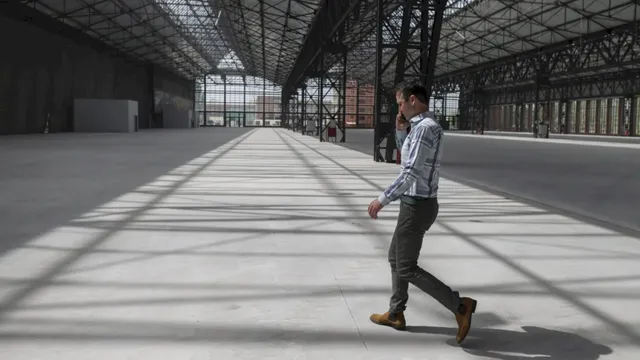 Un bărbat într-un tricou cu mânecă lungă și pantaloni gri merge printr-un spațiu industrial mare și gol cu structură metalică.