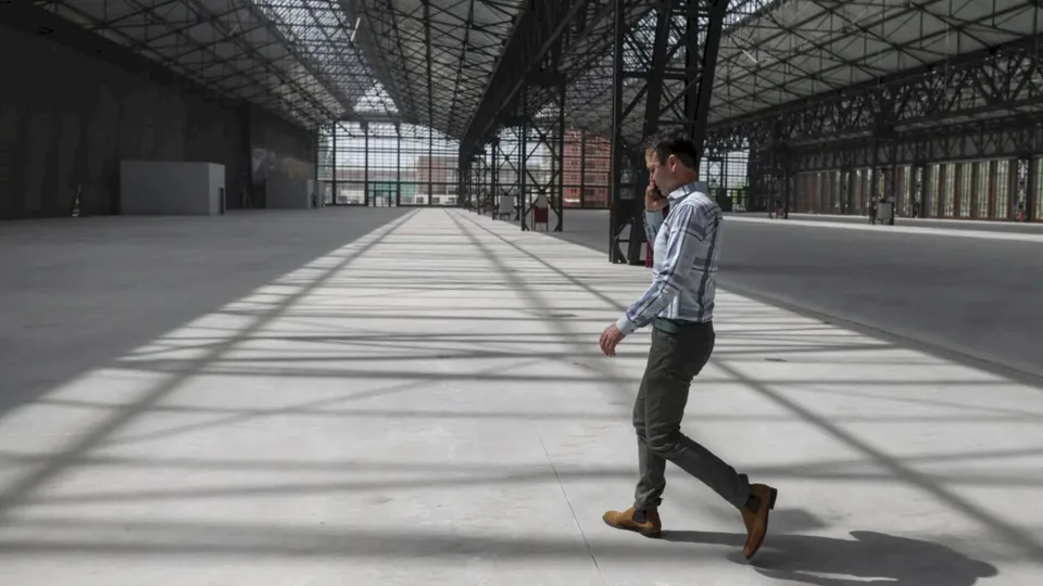 Un bărbat într-un tricou cu mânecă lungă și pantaloni gri merge printr-un spațiu industrial mare și gol cu structură metalică.