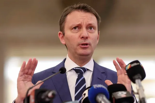 Un bărbat cu cămașă albă, cravată albastră cu dungi și sacou albastru ridică ambele mâini în timp ce vorbește în fața mai multor microfoane.