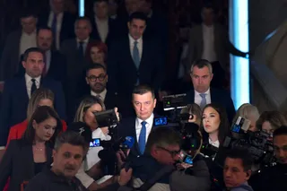 Un bărbat într-un costum albastru cu cravată albastră stă înconjurat de microfoane și camere de filmat în fața unui grup de persoane.