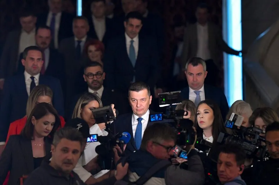 Un bărbat într-un costum albastru cu cravată albastră stă înconjurat de microfoane și camere de filmat în fața unui grup de persoane.