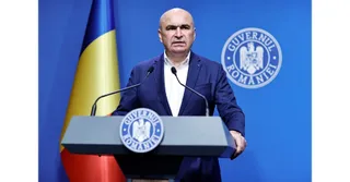 Un bărbat cu părul scurt și sacou albastru stă în spatele unui podium gri cu sigiliul Guvernului României în fața unui fundal albastru.