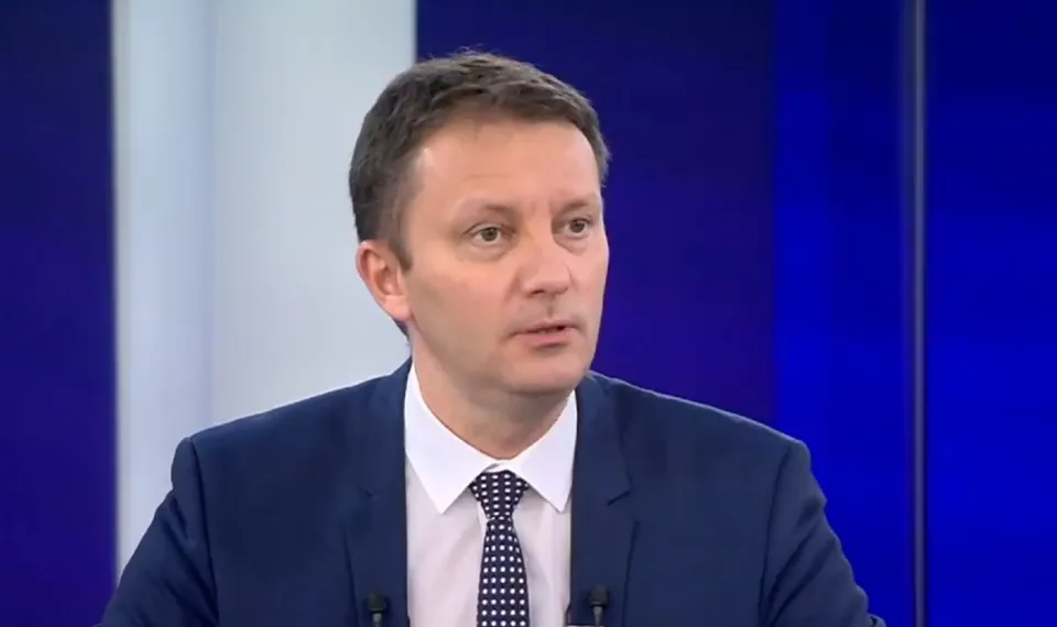 Un bărbat cu părul scurt, purtând o cămașă albă, o cravată cu puncte și un sacou albastru, este poziționat în fața unui fundal albastru.