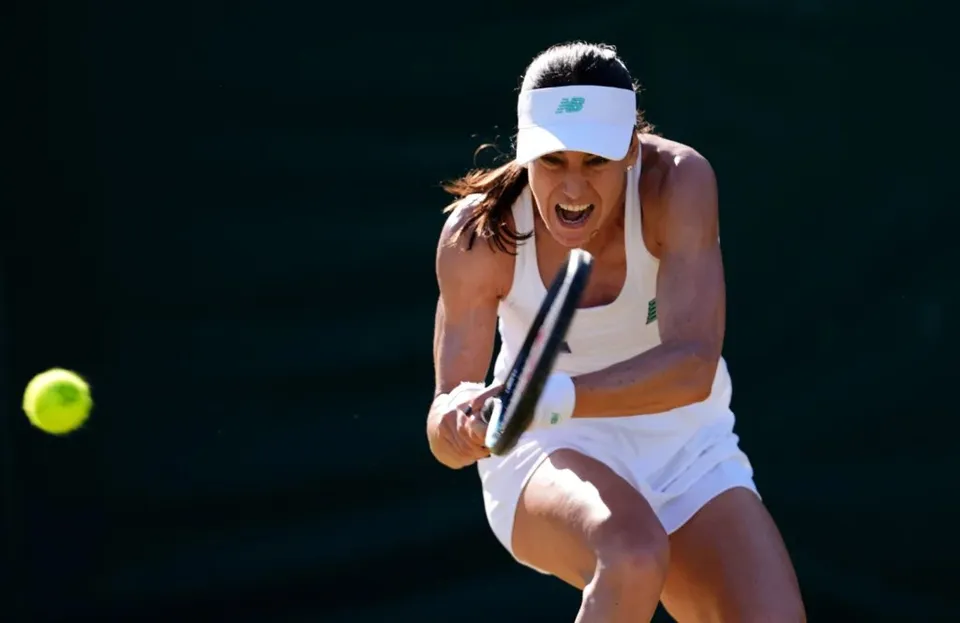 O femeie cu vizieră albă și echipament sportiv alb lovește o minge de tenis cu o racletă pe un teren de culoare închisă.