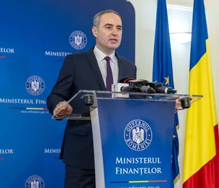 Un bărbat într-un costum închis și cravată purtând o cămașă albă stă în spatele unui podium albastru cu inscripții și microfoane.