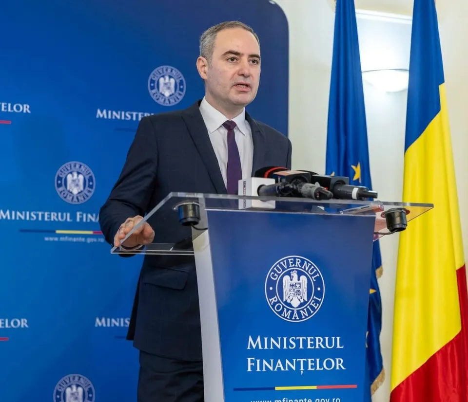 Un bărbat într-un costum închis și cravată purtând o cămașă albă stă în spatele unui podium albastru cu inscripții și microfoane.
