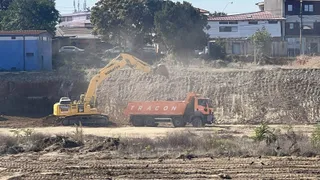 O excavatoare galbenă descarcă pământ într-un camion portocaliu pe un șantier de construcții aflat lângă câteva clădiri și copaci.